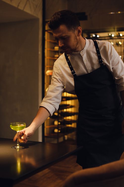 Free A bartender in an apron serving a green cocktail inside a stylish indoor bar setting. Stock Photo