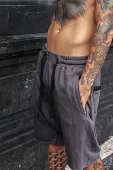 Shirtless tattooed man in gray shorts with hand in pocket, showcasing body art and fitness.