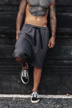 Shirtless tattooed man in urban setting with casual shorts and sneakers.