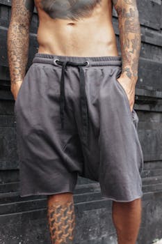 A fit tattooed man stands confidently in gray shorts with hands in pockets.