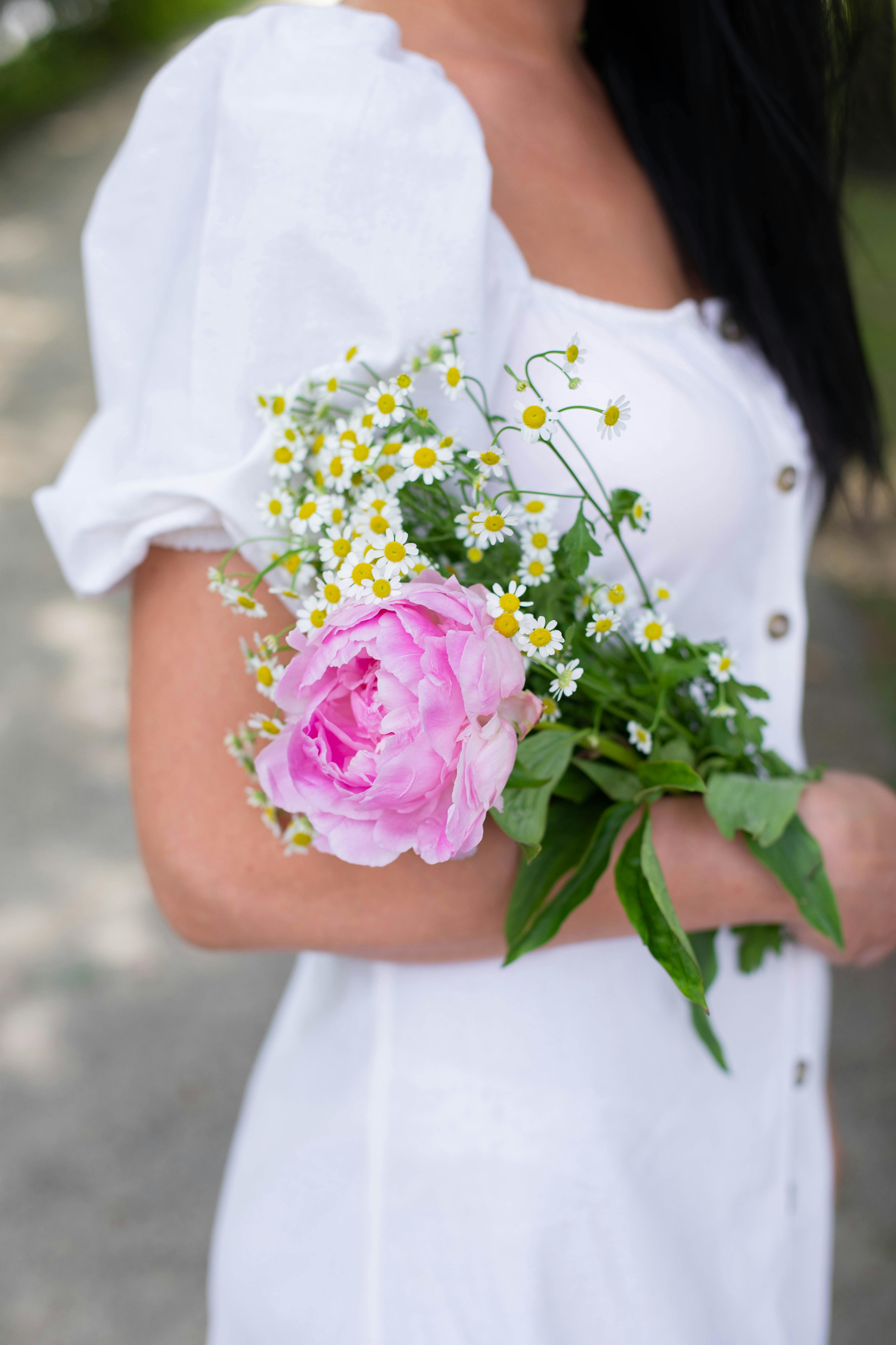 Close-up of a woman in a white dress holding a bouquet with pink peony and chamomile outdoors.