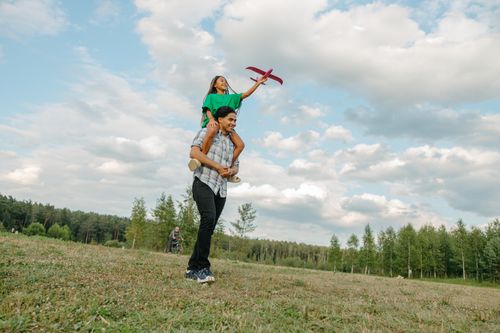 Free A joyful father carrying his daughter on shoulders in a sunny park playing with a toy plane. Stock Photo