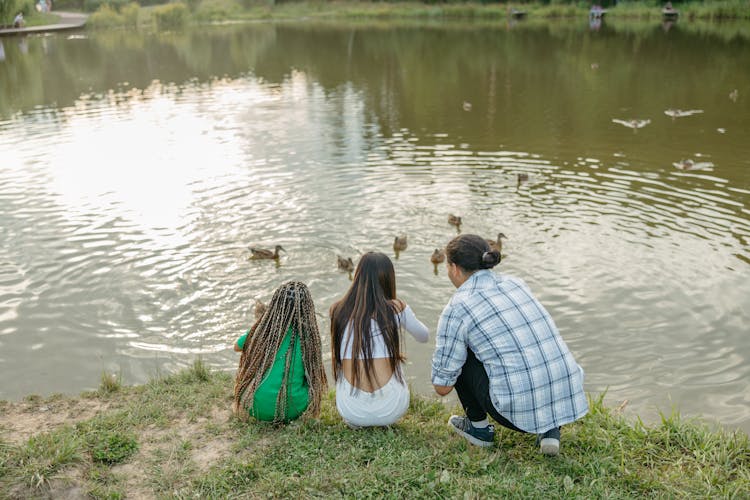 Teenage Boy And Girls Sitting On A Grass In Front Of A Ducks On A Pond