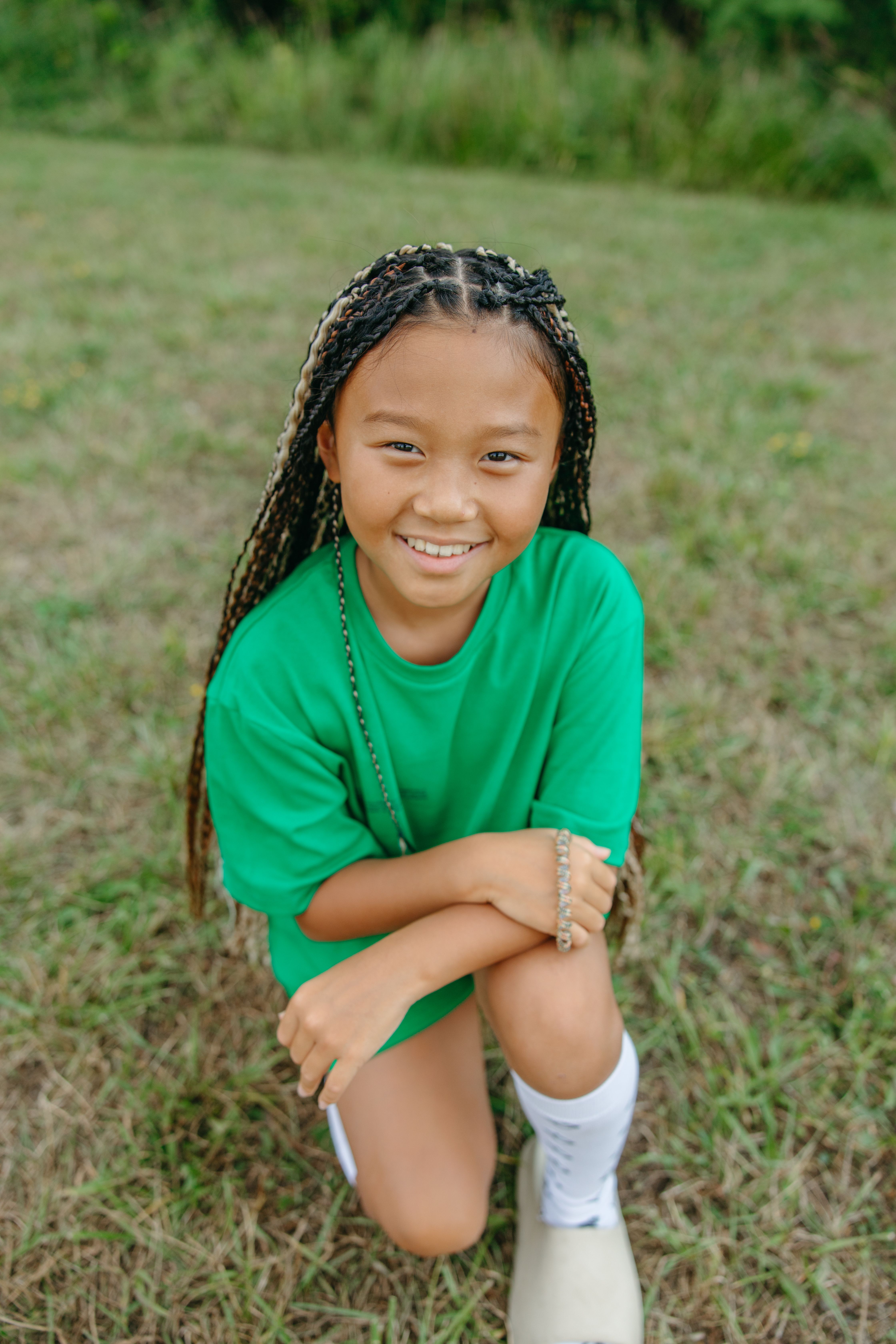 Happy child with braided hair kneeling in a grassy field, wearing a green shirt.