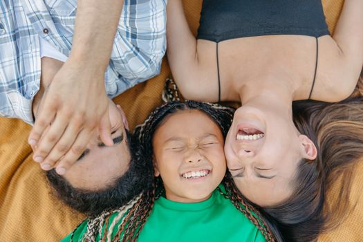 A joyful family bonding moment with parents and daughter laughing while lying on a blanket outdoors.