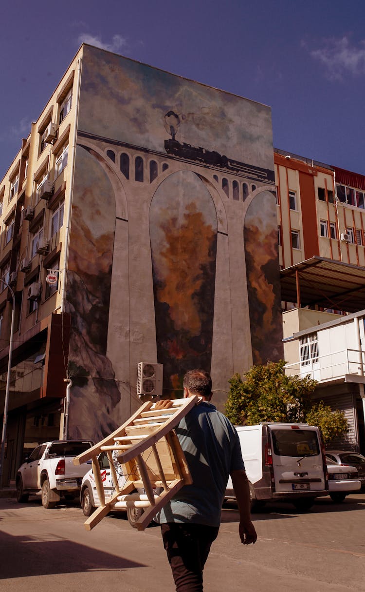 A Man Carrying A Wooden Chair While Walking At The Car Park Near A Building With Painting On The Wall