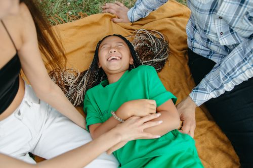 Free Joyful girl laughing while being tickled by family members outdoors on a blanket. Stock Photo