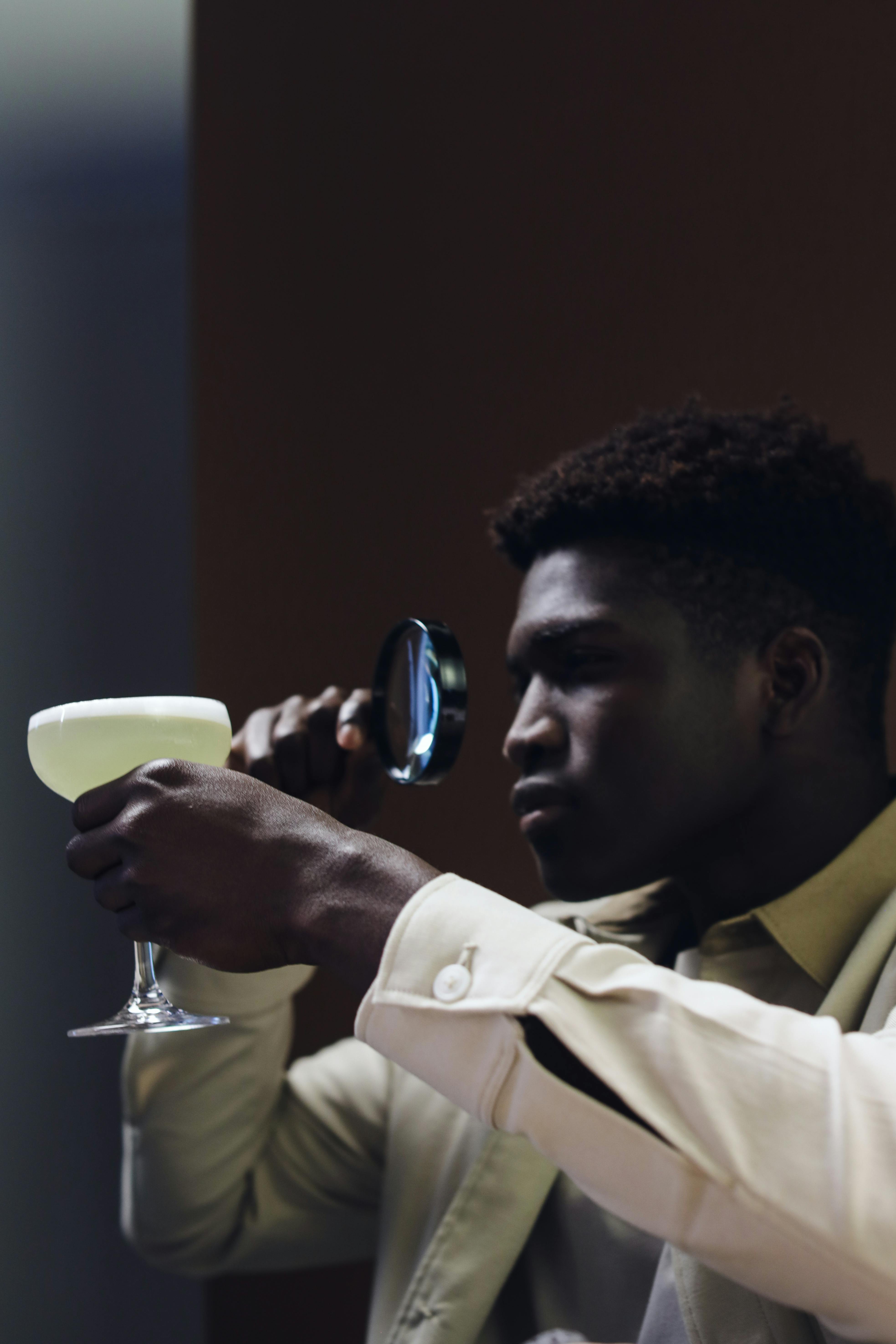 African American man closely examines a cocktail in a glass using a magnifying glass indoors.