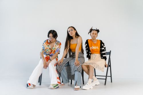 Free Three young models posing on chairs with a stylish and colorful fashion expression against a white studio backdrop. Stock Photo