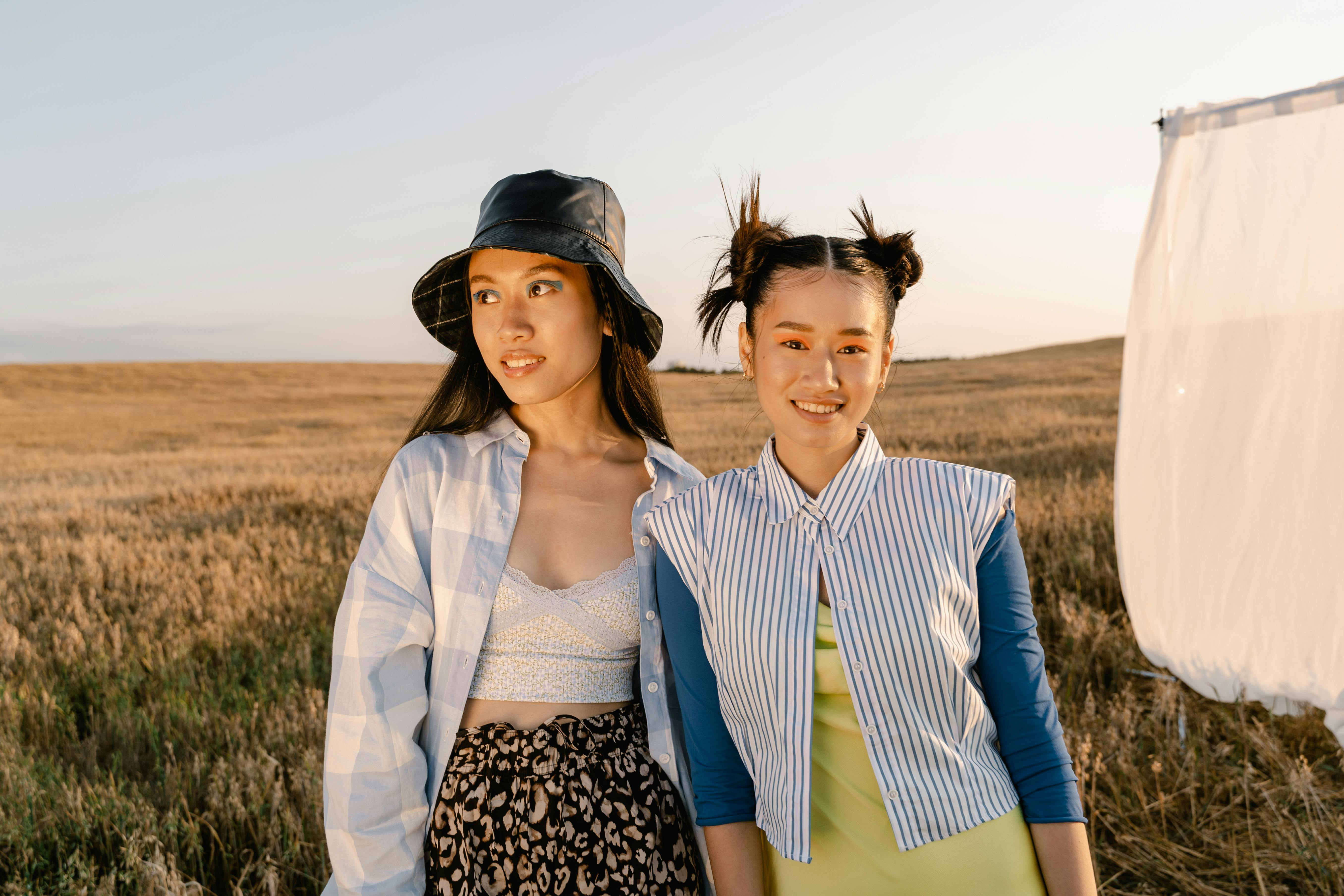 Two fashionable young women pose in a wheat field, showcasing modern style and summer vibes.