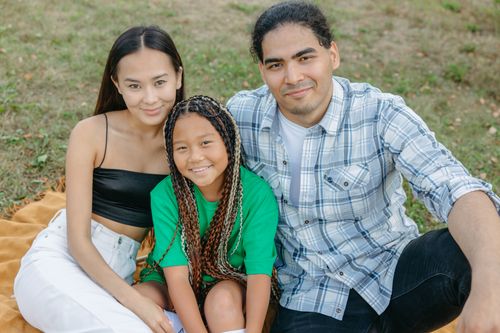 Free A family of three smiling and sitting on a blanket outdoors, enjoying quality time together. Stock Photo