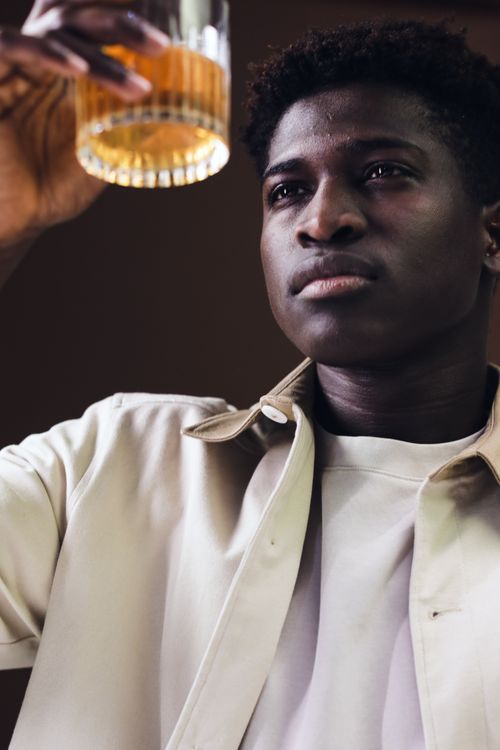 Free A young black man in a white shirt holding a whiskey glass, examining it thoughtfully indoors. Stock Photo