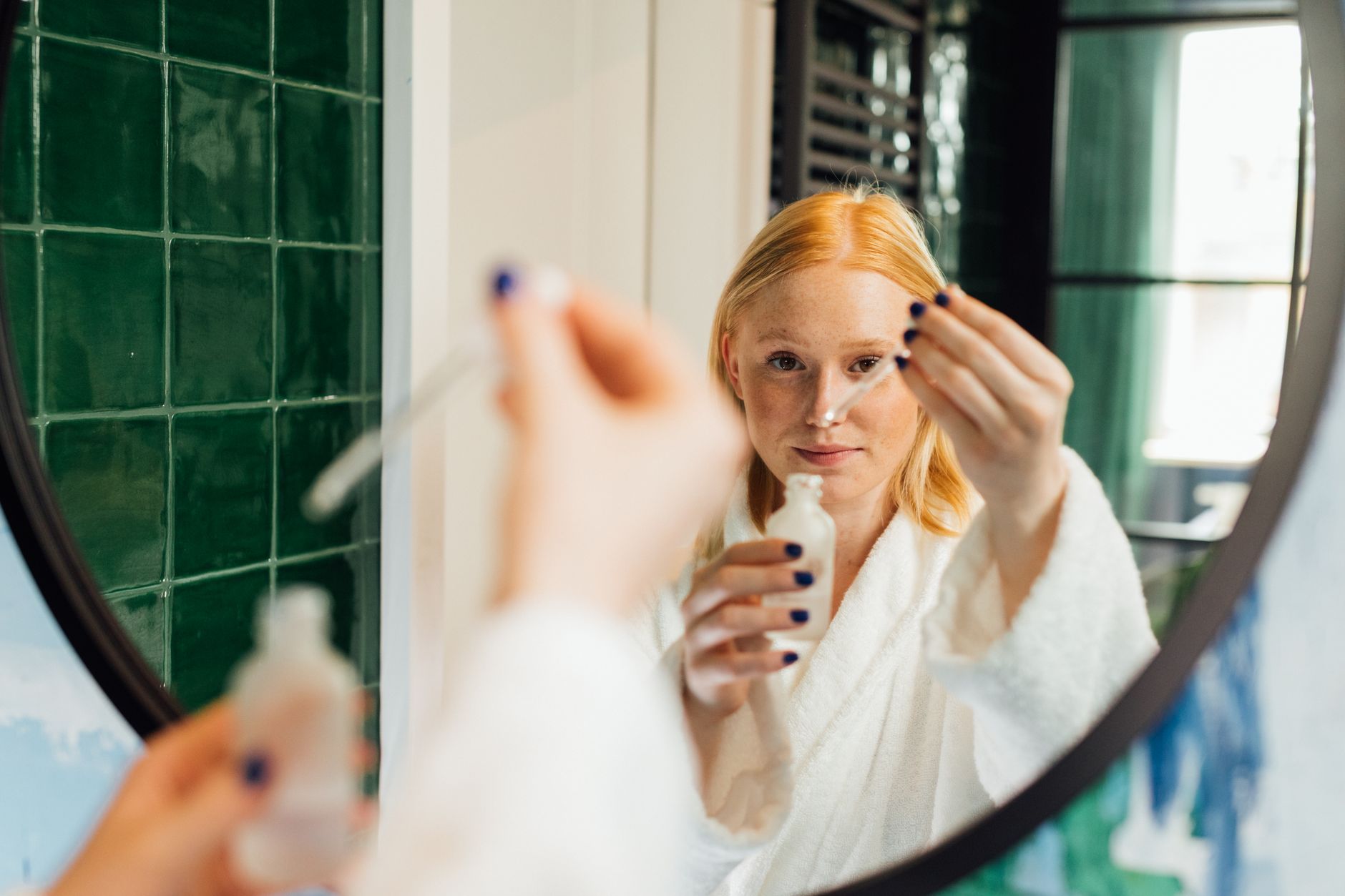 Adult woman applying skincare serum while wearing a bathrobe in front of a bathroom mirror.