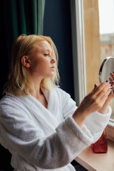 A blonde woman in a white robe holding a mirror by a window, reflecting thoughtful mundanity.