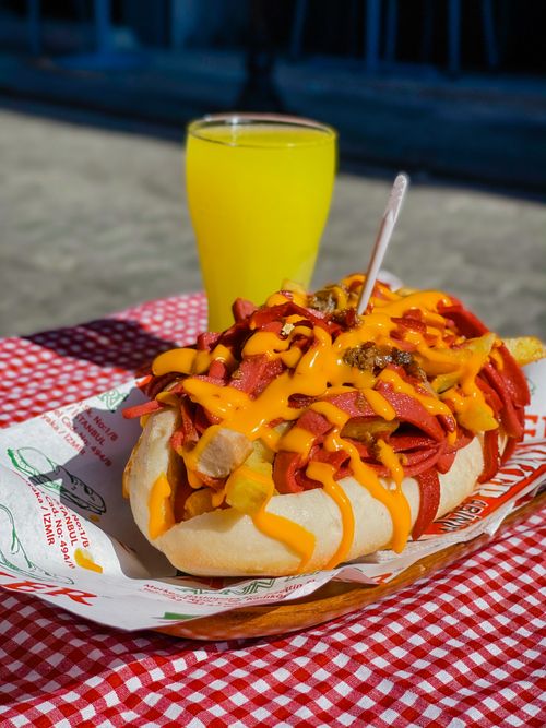 Free Close-up of a loaded hot dog with cheese and a yellow beverage on a picnic table. Stock Photo