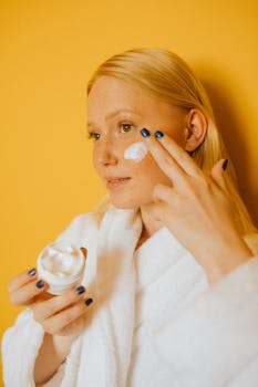 Woman in bathrobe applying skincare cream to face against a vibrant yellow background.