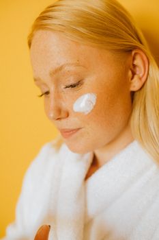 Close-up of a woman in a bathrobe applying cream on her cheek against a yellow background.