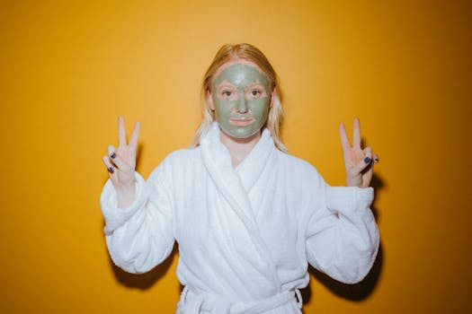 A young woman with a facial mask poses in a bathrobe, making peace signs, against a vibrant yellow background.