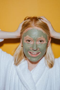 Portrait of a smiling woman in a robe with a green face mask against a yellow background, emphasizing skincare and self-care.