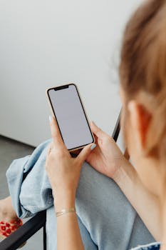 Woman sitting indoors using a smartphone with a blank screen.