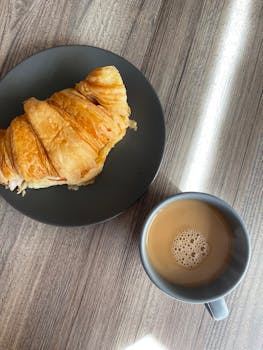 A close-up of a fresh croissant with a cup of coffee on a wooden table. Perfect breakfast vibe.