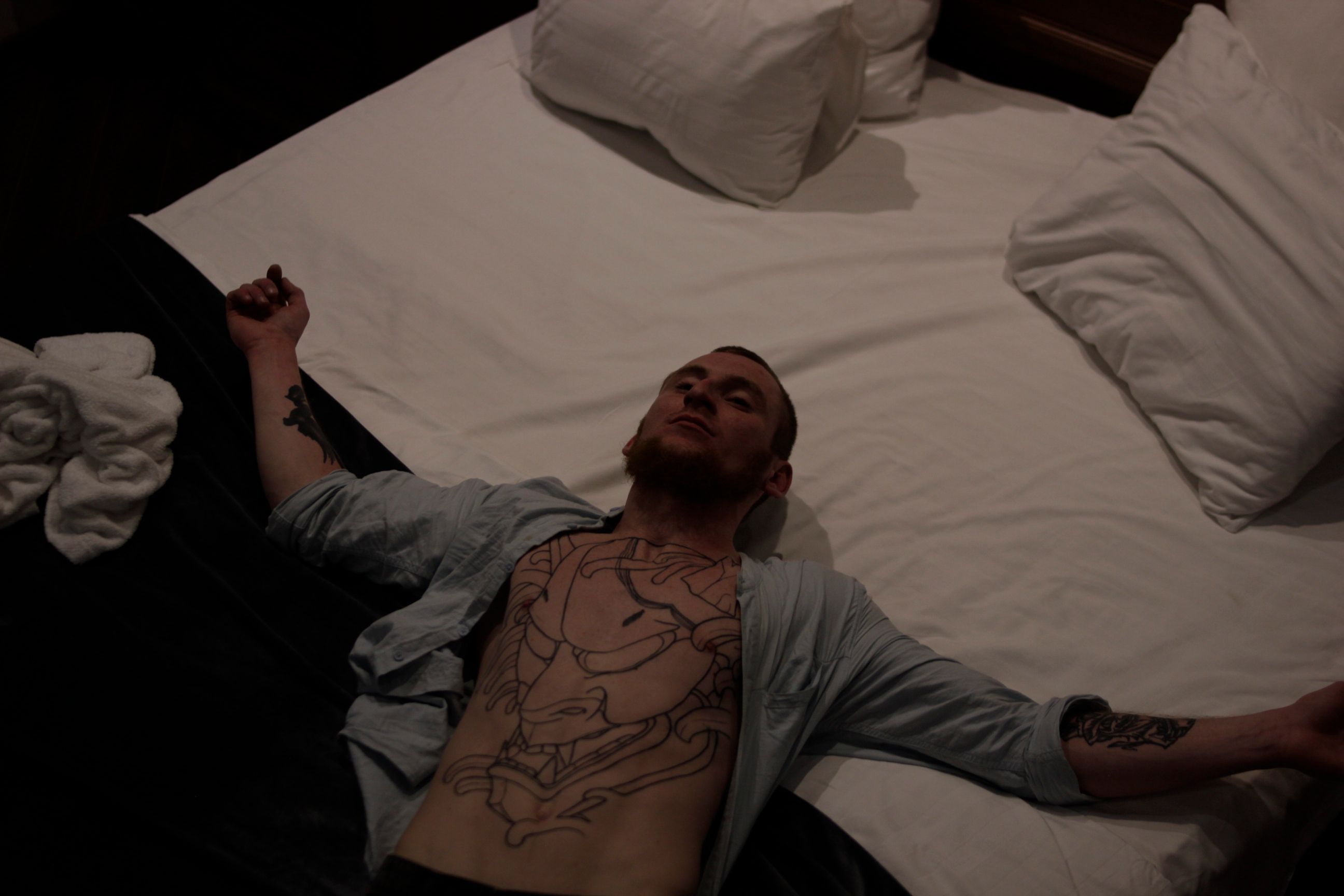 Caucasian man with tattoos lying relaxed on a bed in a dimly lit bedroom.