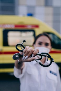 A female paramedic wearing a mask holds a stethoscope with an ambulance in the blurred background.