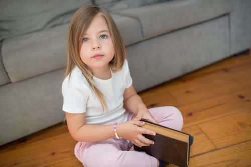 Free Young girl with long hair sitting on a wooden floor, holding a book indoors. Stock Photo