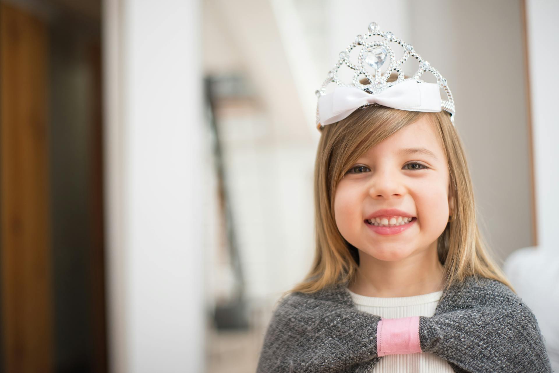 A young girl wearing a decorative crown smiles brightly indoors, embodying playfulness.