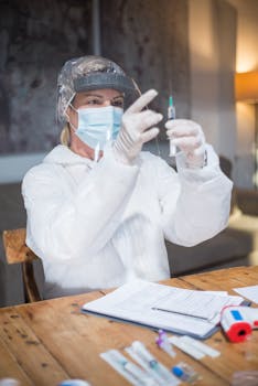 Healthcare worker in PPE preparing a vaccine syringe in an indoor setting.