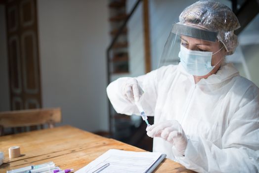 A female healthcare worker in protective gear prepares a syringe at a wooden table indoors.