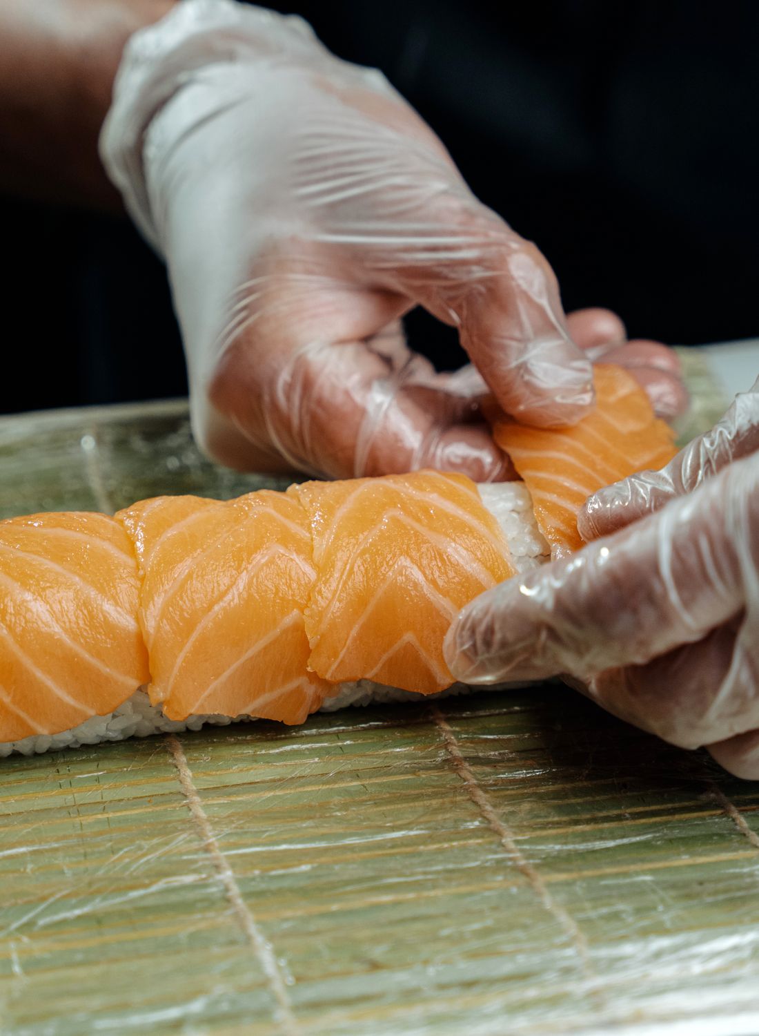 Close-up of chef's hands in gloves crafting salmon sushi roll, showcasing sushi preparation.