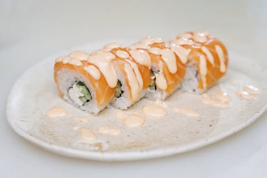 Close-up of a salmon sushi roll topped with creamy dressing, presented on a white plate.