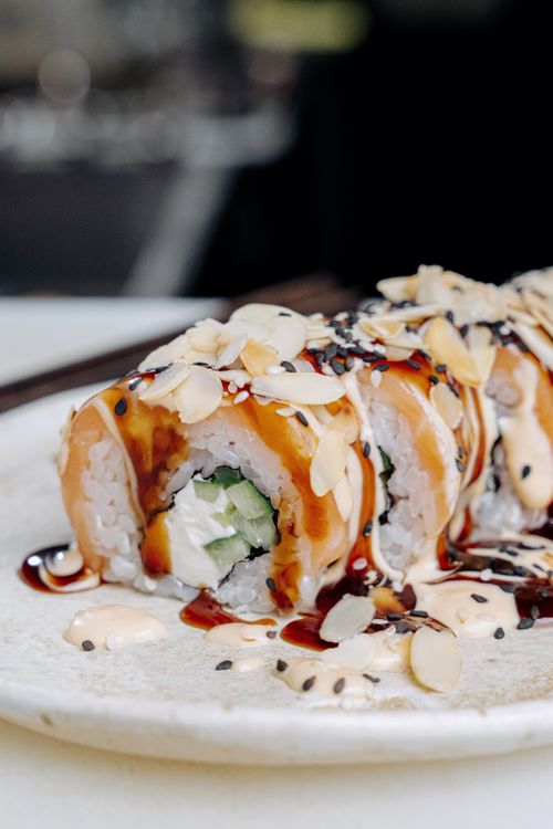 Free Appetizing sushi roll topped with almond flakes and drizzled sauce on a white plate. Stock Photo