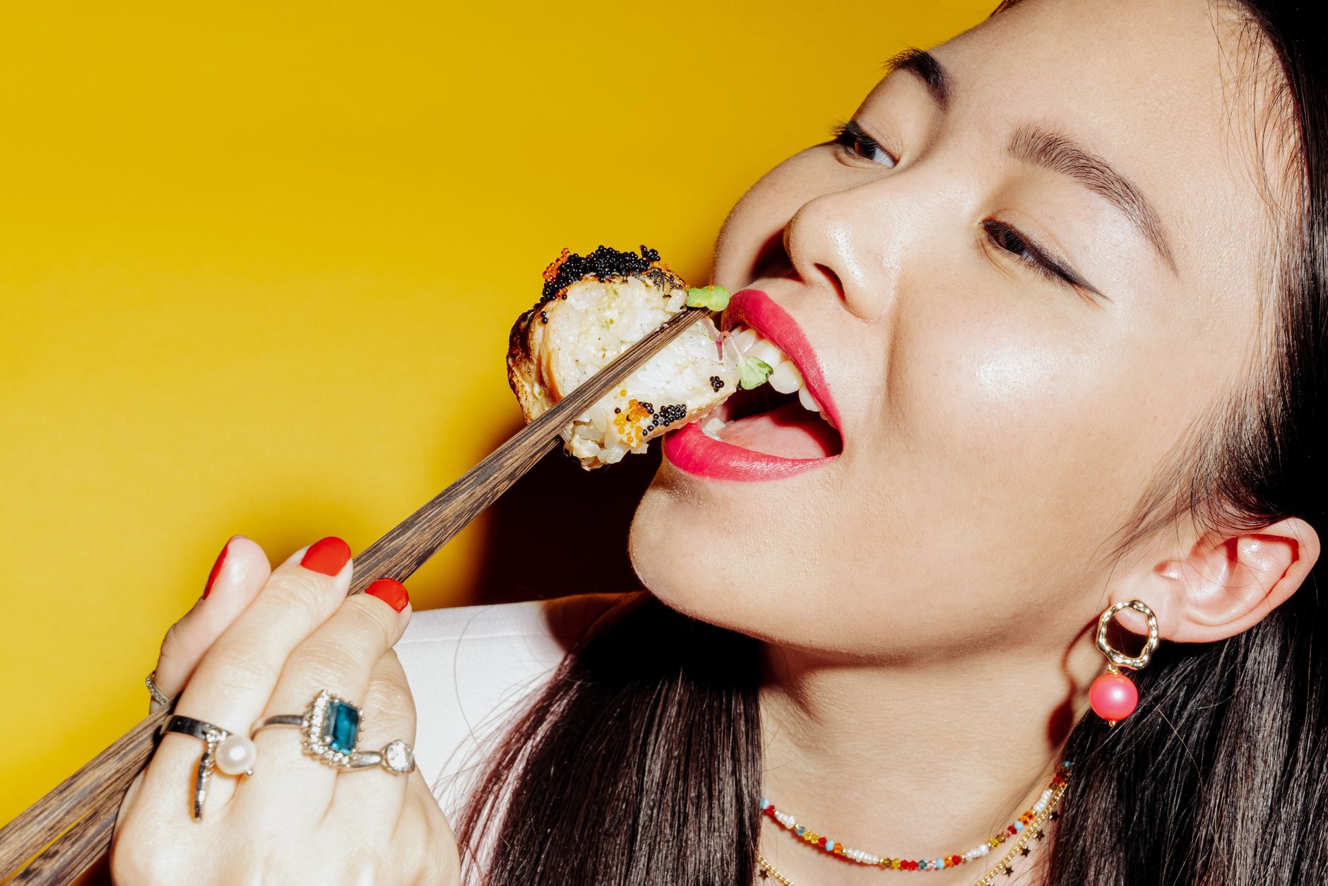 A stylish Asian woman savoring sushi with chopsticks against a vibrant yellow backdrop.