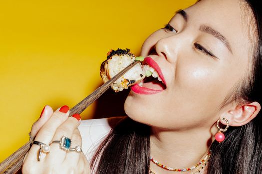 A stylish Asian woman savoring sushi with chopsticks against a vibrant yellow backdrop.