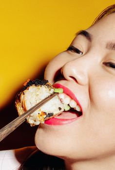 Asian woman savoring a sushi roll with chopsticks against a vibrant yellow background.