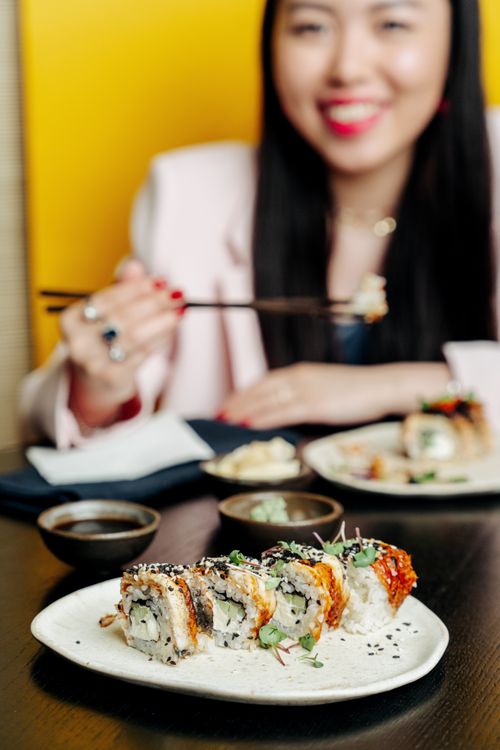 Free Close-up of sushi rolls with a smiling Asian woman in background holding chopsticks. Stock Photo