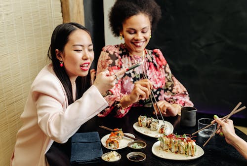 Free Two women smiling and taking photos while enjoying sushi at a Japanese restaurant. Stock Photo
