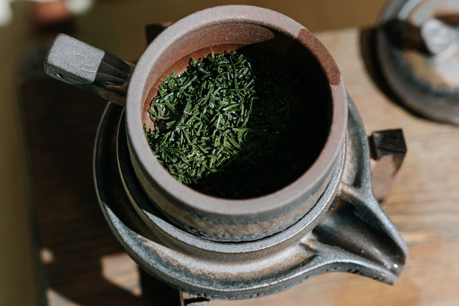 Close-up of a rustic clay teapot filled with dry green tea leaves, highlighting its texture and detail.