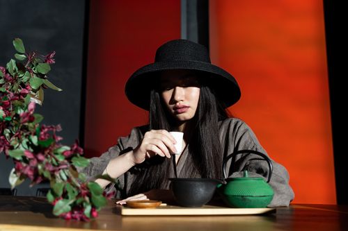 Free Asian woman in a hat enjoying a traditional tea ceremony indoors with floral decor. Stock Photo