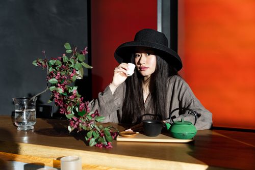 Free Asian woman with black hat sipping tea at a table with vibrant floral decor. Fashionable and serene ambiance. Stock Photo