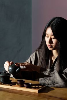 Serene Asian woman performing a traditional tea ceremony with elegant teapot and cups in a sunlit room.