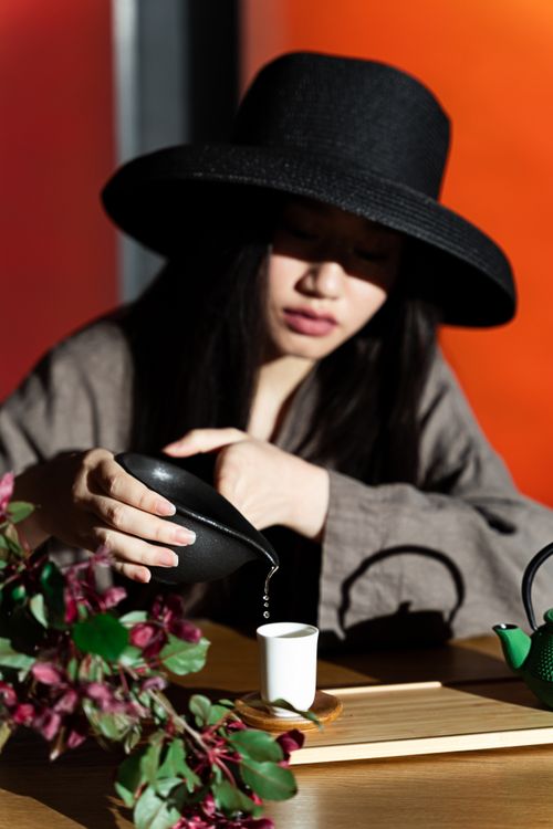 Free A woman in a stylish hat pouring tea from a black teapot into a white cup, bathed in natural sunlight. Stock Photo