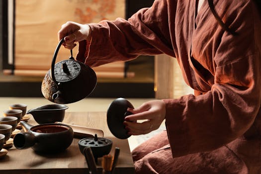 Person in robe performing traditional tea ceremony with ceramic teapots and cups.