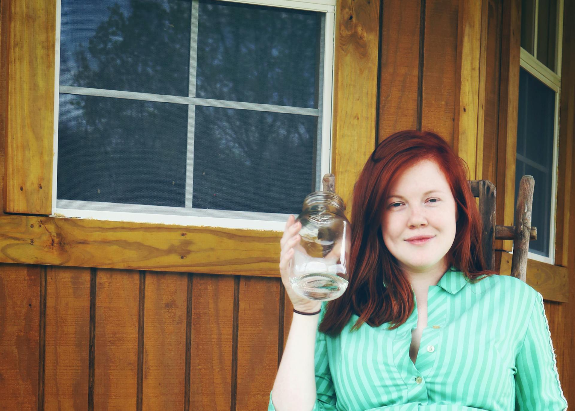 Smiling young woman holding a glass jar on a wooden porch in the countryside.