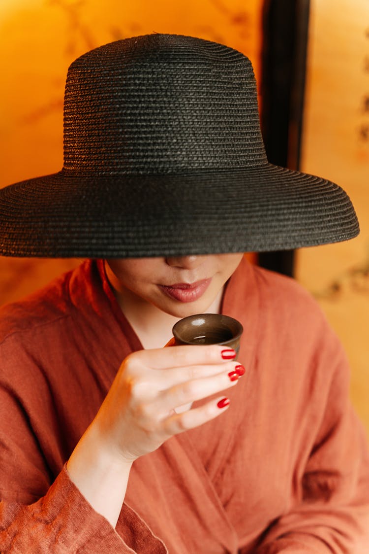 A Woman In Robe And Black Hat Drinking Tea