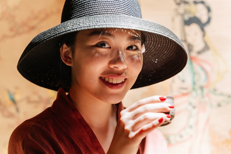 A Smiling Woman Wearing A Black Hat While Holding A Teacup