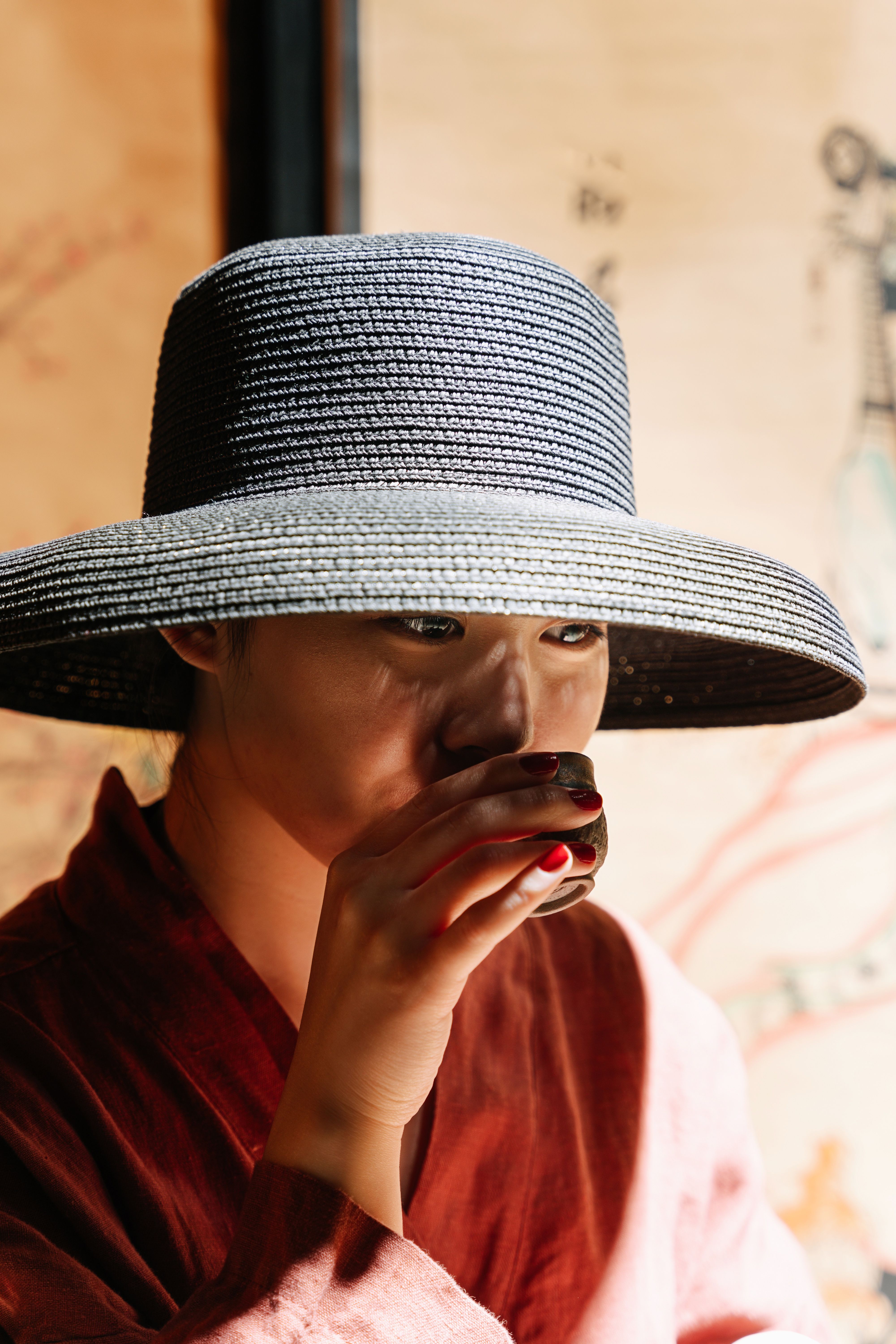 Asian woman enjoying a traditional tea ceremony indoors, wearing a large hat and sipping tea.