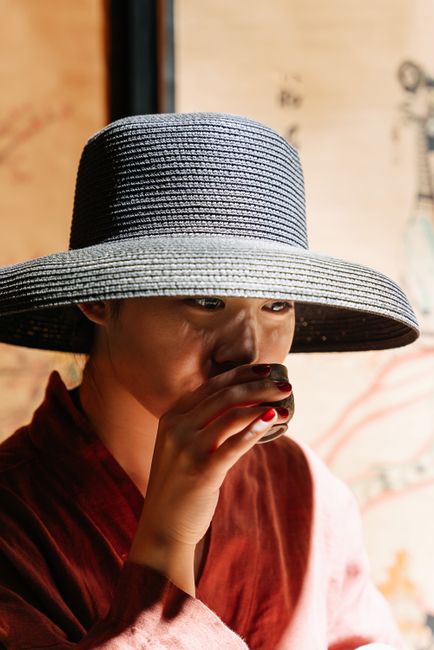 Asian woman enjoying a traditional tea ceremony indoors, wearing a large hat and sipping tea.
